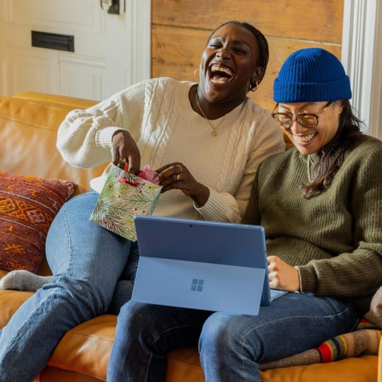 twee lachende mensen op een bank, een van de twee heeft een laptop op schoot